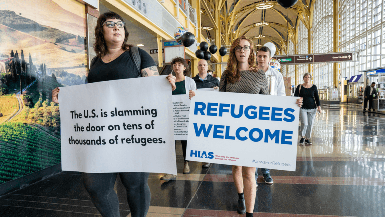 A demonstration at Reagan Airport in DC. Participants hold signs; one reads "The U.S. is slamming the door on tens of thousands of refugees." Another reads "Refugees Welcome" with the HIAS logo and hashtag "Jews for Refugees".