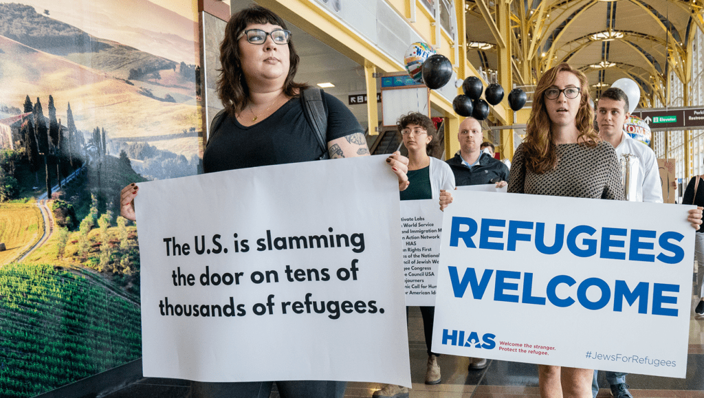 A demonstration at Reagan Airport in DC. Participants hold signs; one reads "The U.S. is slamming the door on tens of thousands of refugees." Another reads "Refugees Welcome" with the HIAS logo and hashtag "Jews for Refugees".