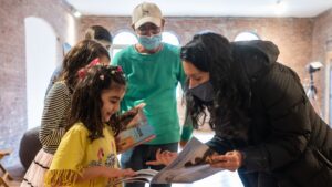 Zubair Babakarkhail, center, watches as his daughter Javeria, left, 6, points out photos of other refugees she knows in an issue of “Postindustrial” magazine to Ivonne Smith-Tapia, right, director of refugee services at Jewish Child and Family Service (JCFS), during a small gathering on Sunday, Dec. 19, 2021, in Pittsburgh. Babakarkhail is continuing reporting on his home country of Afghanistan for the magazine and other news outlets after his role as a journalist put him in danger under Afghanistan’s new Taliban rule. (Stephanie Strasburg/HIAS)
