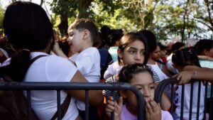 Group of migrant women and children standing behind a metal barricade, looking somber while waiting outdoors near the U.S.-Mexico border.