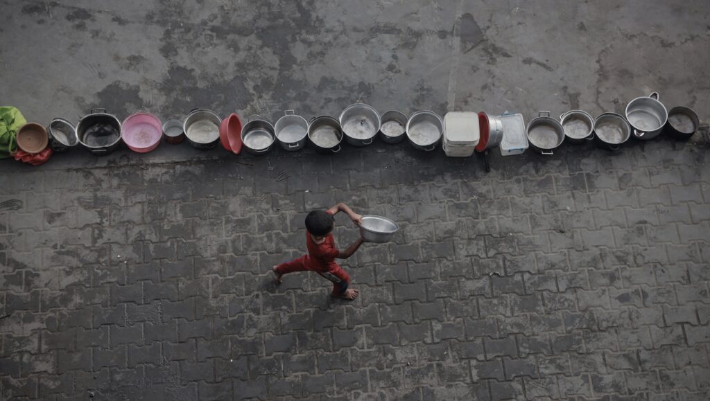 A little boy runs with an empty pail in search of food beside a column of other empty pails.