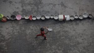 A little boy runs with an empty pail in search of food beside a column of other empty pails.