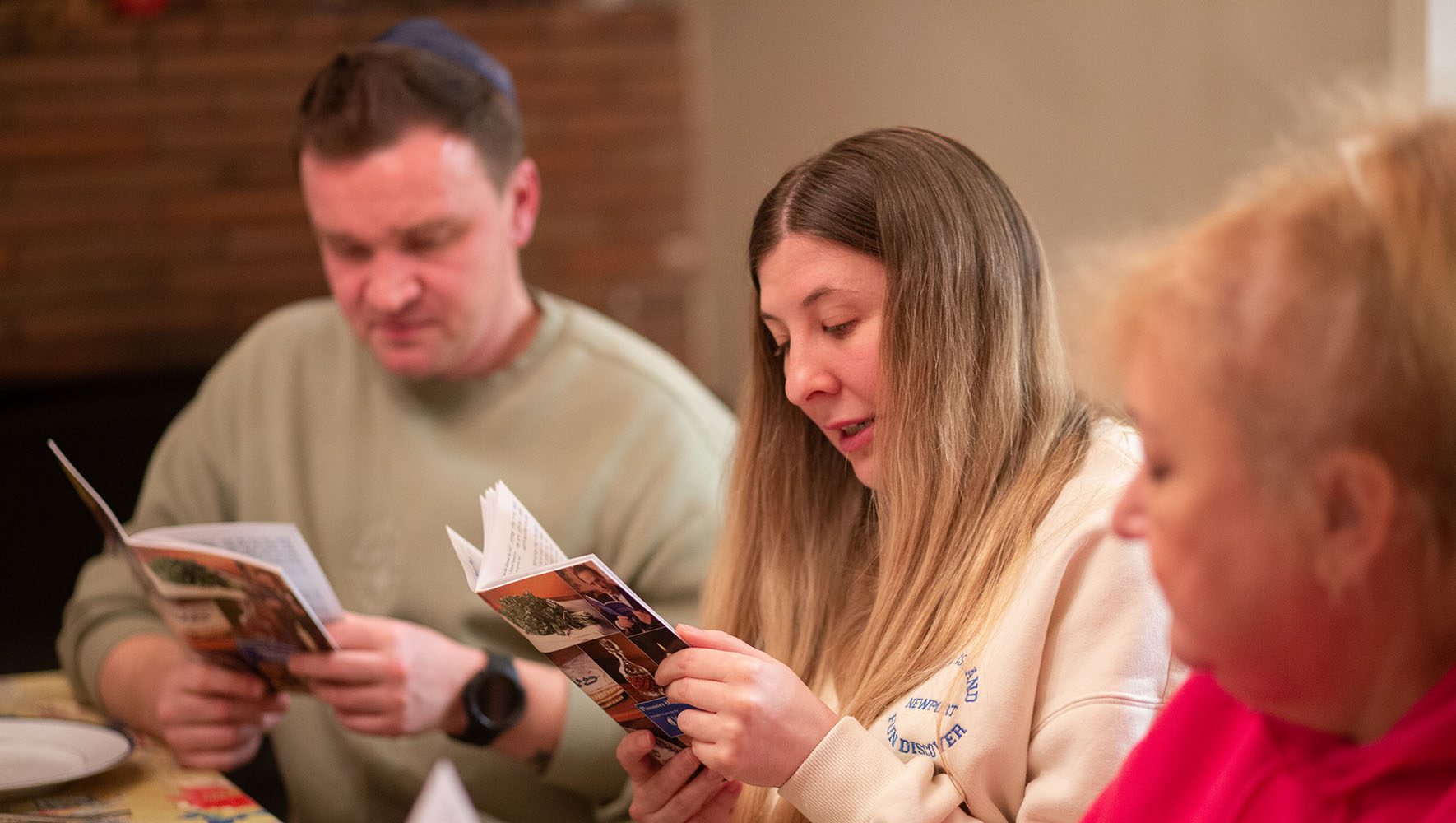 Daria Levit, center, reads from the Haggadah in English as her husband Eduard Levit,left, and mother-in-law Tamila Kushnarova, right, listen, during the Passover Seder. | In Time for Passover, Ukrainians Find Jewish Community in U.S. | HIAS