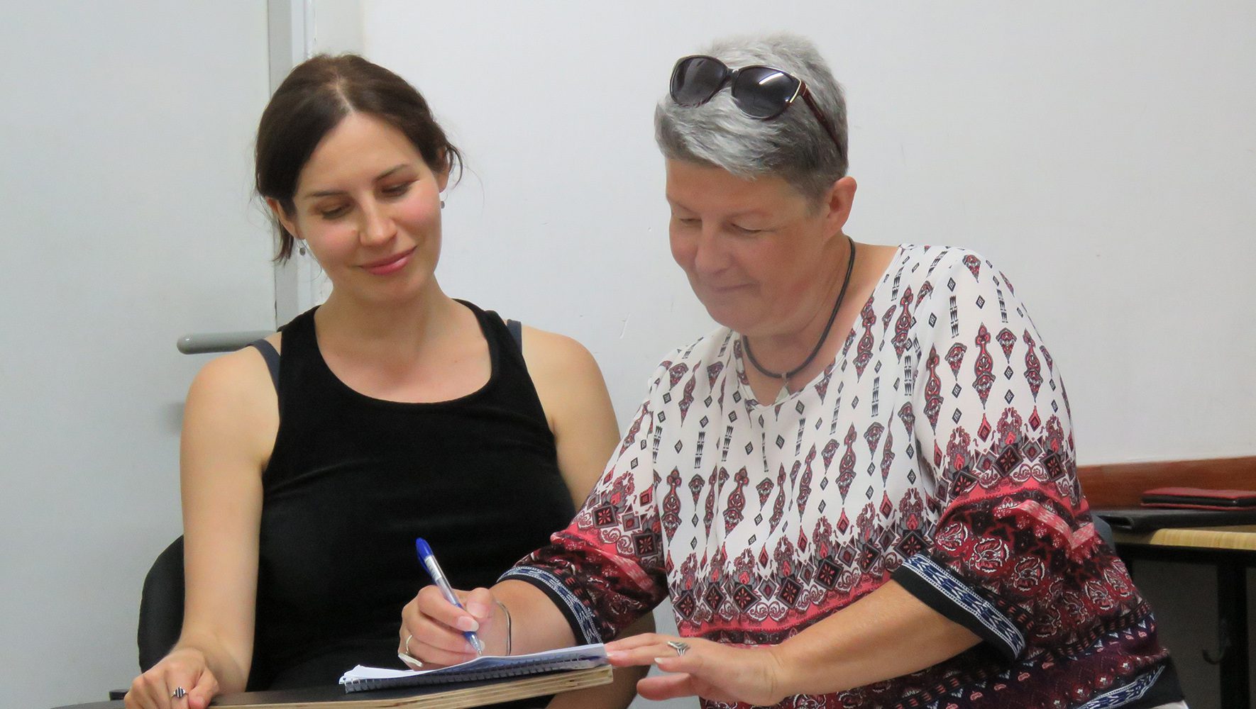 Israeli woman smiles as Ukrainian woman sits next to her and leans over to write something on a small desk