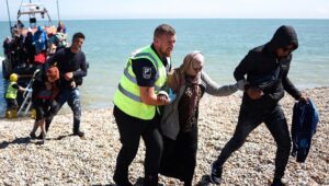An interforce officer helps a woman on the beach at Dungeness on the southeast coast of England after she disembarked from a Royal National Lifeboat Institution (RNLI) lifeboat after being picked up at sea with other migrants while attempting to cross the English Channel from France on August 16, 2023. (Henry Nicholls/AFP via Getty Images)