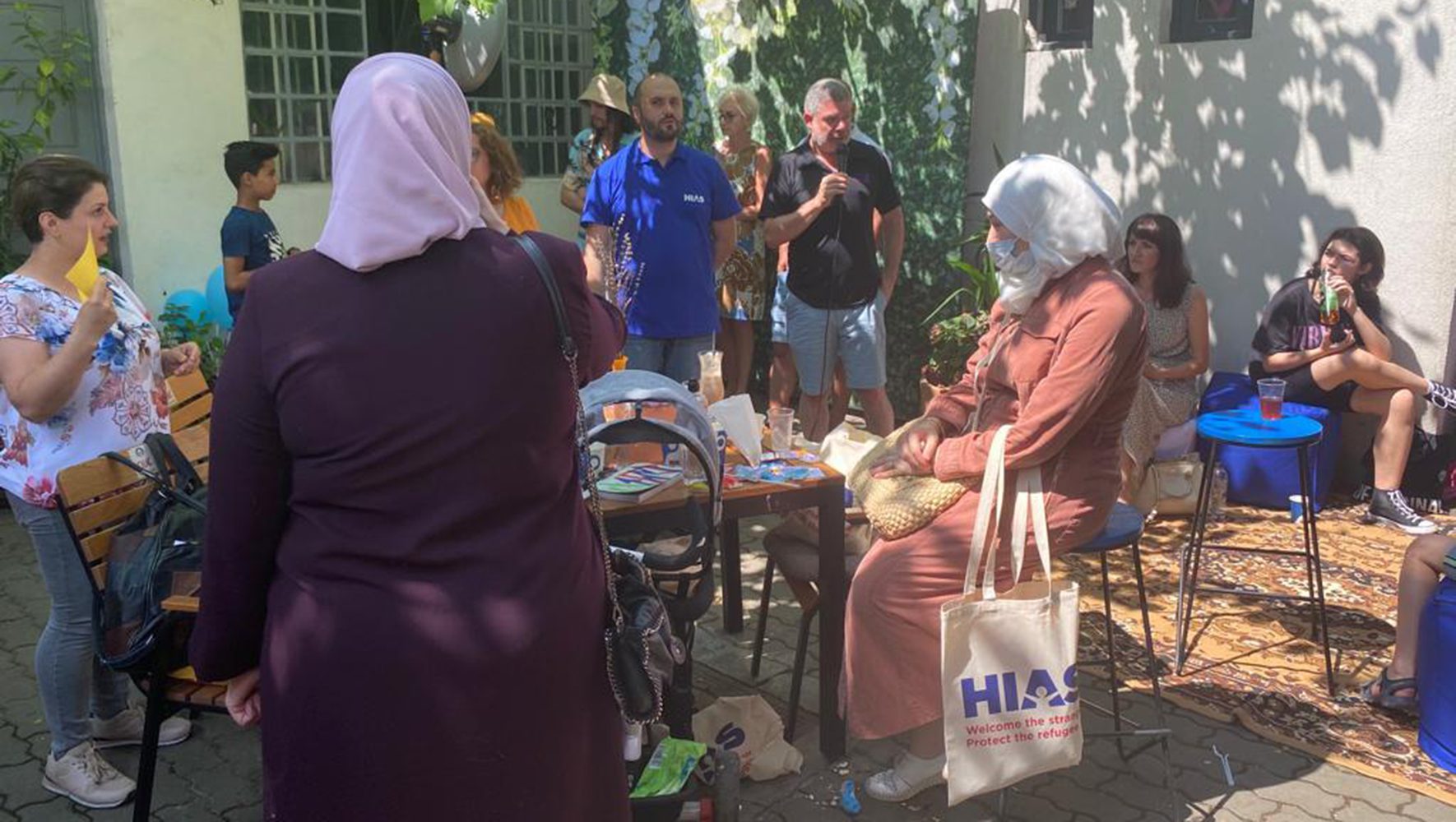 Outside a man talking on microphone, woman in headdress sitting with HIAS tote bag